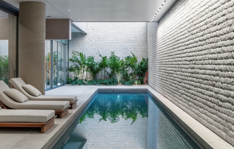 Piscina coberta do Casa Leopoldo, com espreguiçadeiras, parede texturizada e jardim, no Itaim Bibi