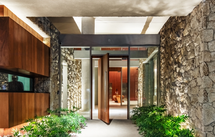 Acesso principal do Casa Leopoldo, com porta pivotante de madeira entre paredes de pedra natural e jardim, no Itaim Bibi